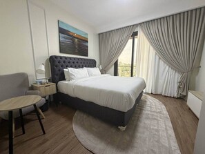 A hotel room with a bed, a desk, a chair, a nightstand, a lamp, and a painting on the wall. - Harmonia Studio | Airport City • Pool • Fast WiFi (Accra)