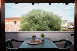 Ein Balkon mit Tisch, Stühlen, einer Kaffeekanne, einem kleinen Topfpflanzen und Blick auf Gebäude und Bäume.