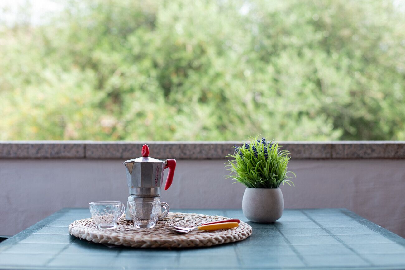 Une table avec une machine à café, deux verres, une cuillère et une plante en pot, le tout posé sur un set de table en fibres tressées.