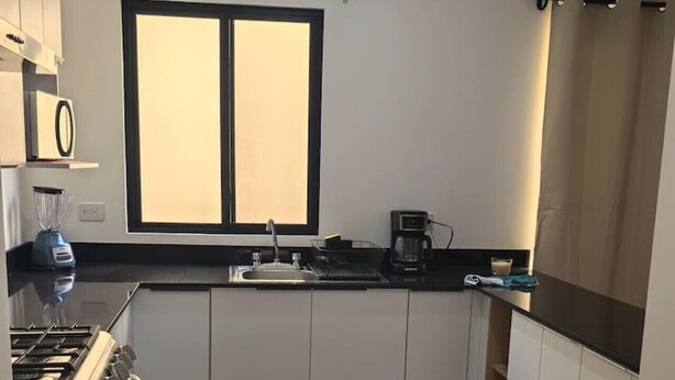 A modern kitchen with white cabinets, a stove, a window with blinds, and a coffee maker. - Vista Central Park Caucel3 Habitaciones (Caucel)