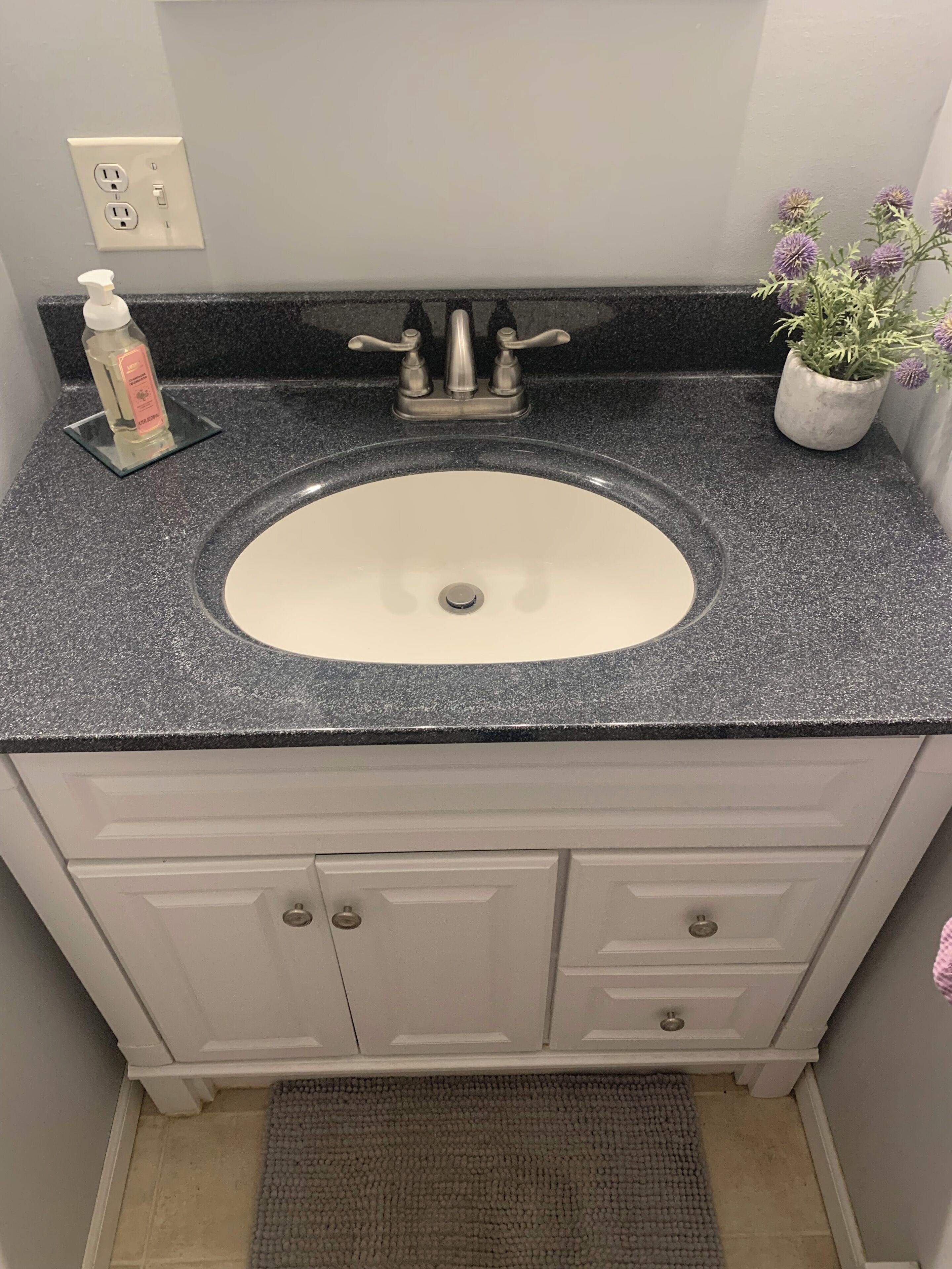 Un baño con lavamanos blanco, encimera de granito oscuro, una planta en maceta y una botella de jabón de manos.