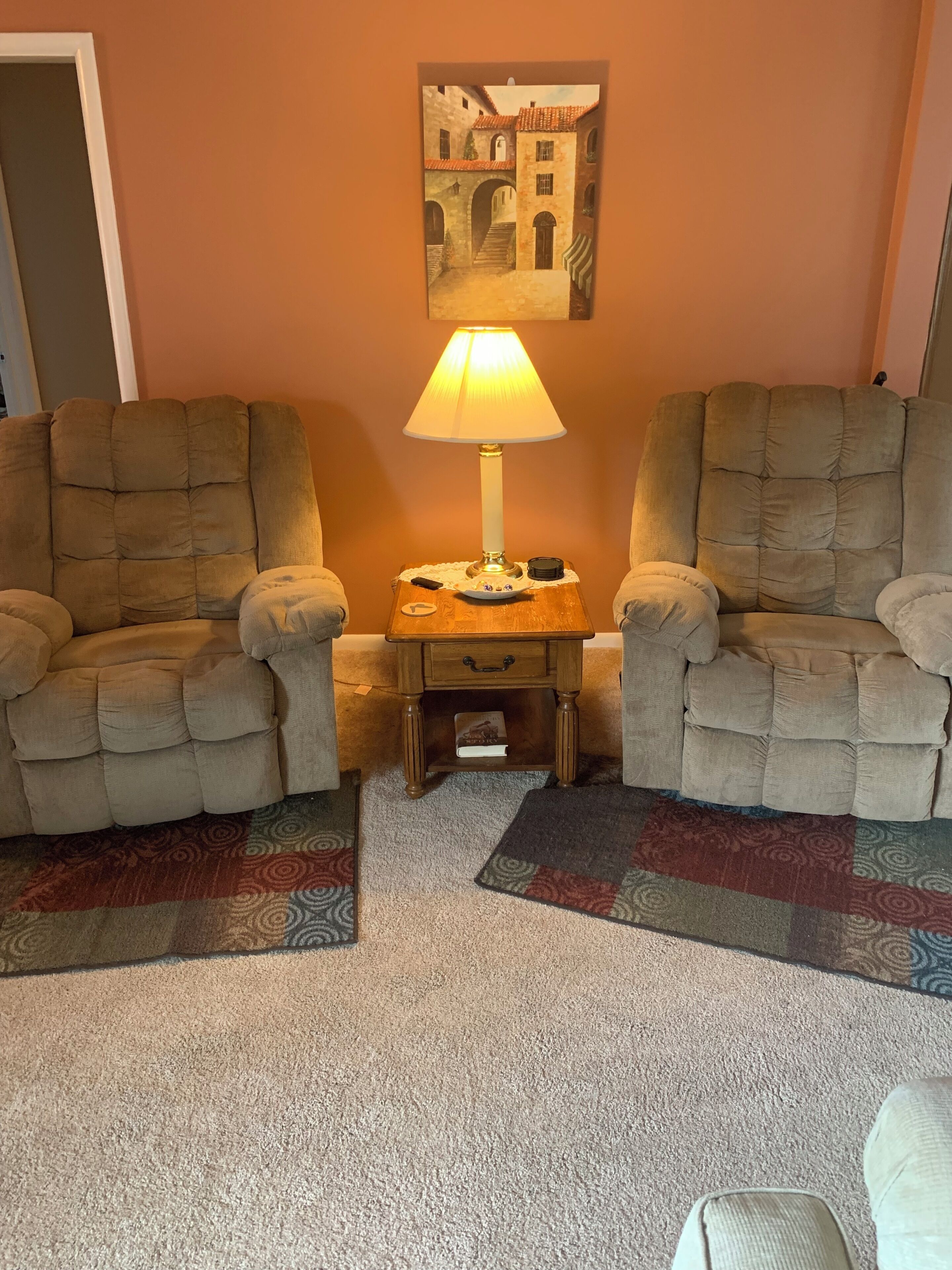 A living room with two armchairs, a small wooden side table with a lamp, a rug, and a painting on the wall.