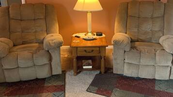 A living room with two armchairs, a small wooden side table with a lamp, a rug, and a painting on the wall.