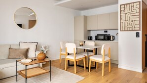 Un salon moderne comprenant un canapé, une table basse et des fauteuils, un miroir rond, ainsi qu’un coin cuisine avec des placards et des appareils électroménagers.