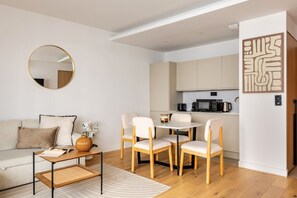 A modern living room with a sofa, coffee table, and chairs, a round mirror, and a kitchen area with cabinets and appliances. - 2-room apartment - exceptional terrace – Heart of the Presqu'île (Lyon)