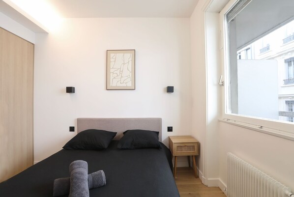 Un dormitorio con cama, dos mesitas de noche, una lámpara de pared, ventana con vistas y un cuadro enmarcado.