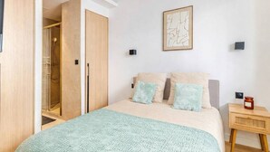 Une chambre à coucher comprenant un lit, une table de chevet, une lampe fixée au mur et une porte donnant accès à une douche.