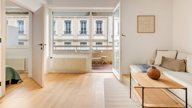 Un appartement moderne avec un plancher en bois, un canapé, une petite table et une vue sur un balcon équipé d’une chaise.