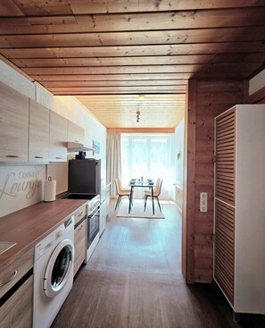 A compact kitchen with wooden cabinets, a washing machine, a dining area with a table and chairs, and a view through a window.