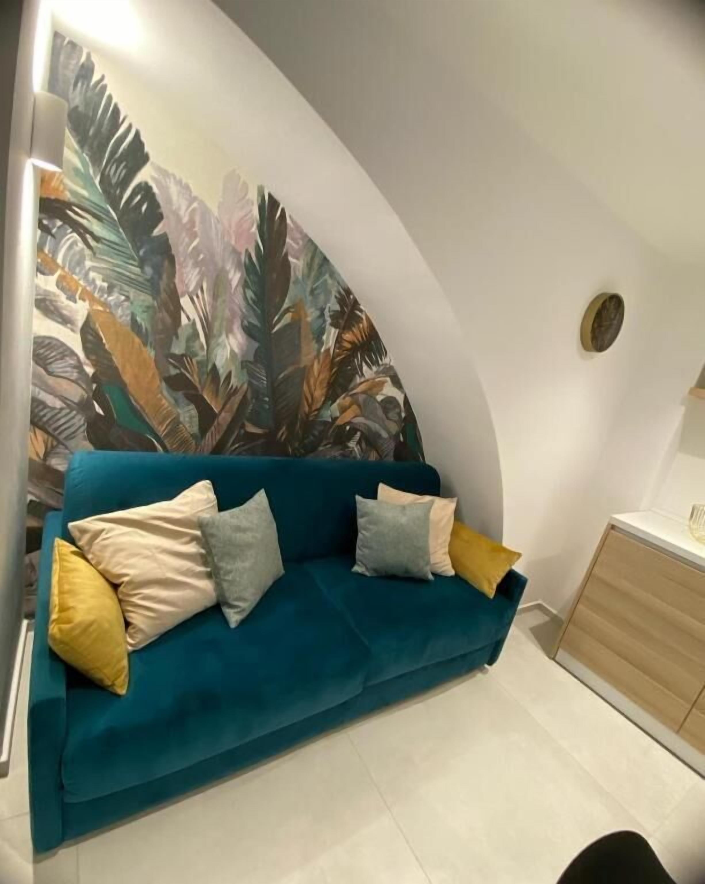 A teal sofa with cushions, a wall with a leaf pattern, and a wooden cabinet.