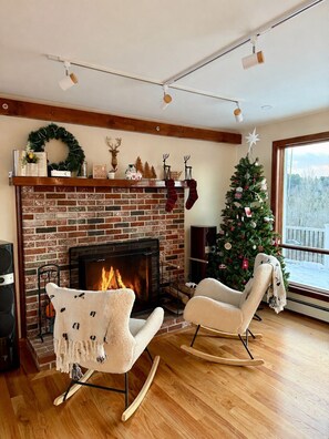 Un salon chaleureux avec une cheminée en briques, deux fauteuils à bascule, un sapin de Noël et une vue sur un paysage enneigé.