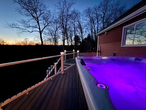 Una terraza de madera que conduce a un jacuzzi con iluminación morada, rodeado de árboles y una casa.