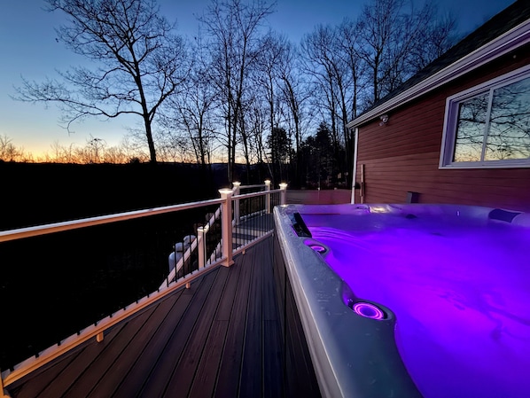 Une terrasse en bois mène à un jacuzzi éclairé de lumières violettes, entouré d’arbres et d’une maison.