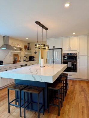 Une cuisine moderne dotée d’un îlot en marbre, de luminaires suspendus, d’un four encastré et de meubles de rangement blancs.