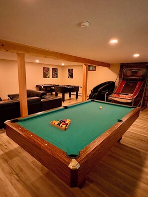 Una mesa de billar con superficie de fieltro verde y un juego de bolas en la esquina, una mesa de fútbol de mesa y una mesa de billar en una habitación con suelo de madera y pared de ladrillo.