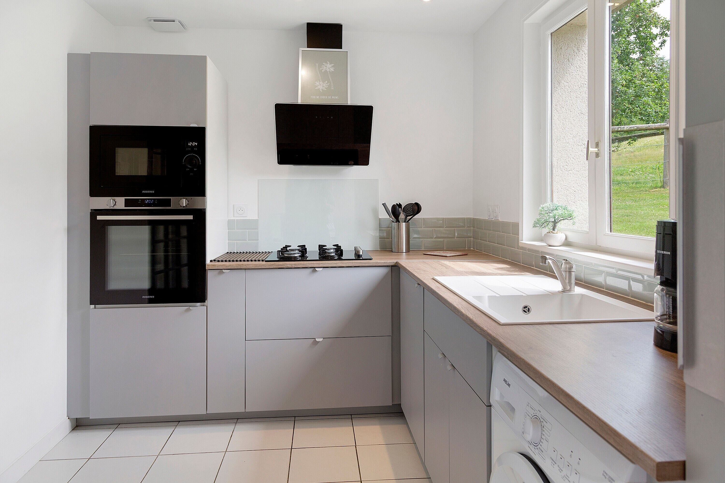 A modern kitchen with a built-in oven, microwave, and dishwasher, a large window with a view of greenery, and a wooden countertop.