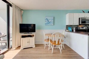Quarto com televisão, mesa de jantar, cadeiras, cozinha e porta de vidro deslizante.