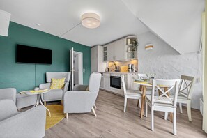 A modern living room with a TV, grey sofas, a dining area with white chairs, and a kitchen with white cabinets. - Kronsgaard Beach Loft - Ocean View, Activity Room, Playground, Balcony (Kronsgaard)