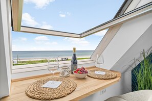 A dining area with a view of the beach, a bottle of wine, and a bowl of fruit. - Kronsgaard Beach Loft - Ocean View, Activity Room, Playground, Balcony (Kronsgaard)