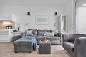 Ein modernes Wohnzimmer mit einem grauen Ecksofa, einem Couchtisch und einem grauen Sessel. An der Wand hängt ein gerahmtes Kunstwerk, im Hintergrund ist eine Küchenzeile zu sehen.