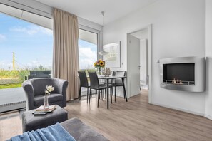 A modern living room with a sofa, a dining table with chairs, a fireplace, and a large window with curtains. - Spacious Apartment with exceptional Sea View and Wellness Area! (Wendtorf)