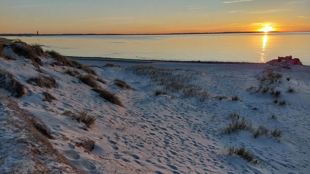 En strand med sanddyner och en solnedgång över vattnet.