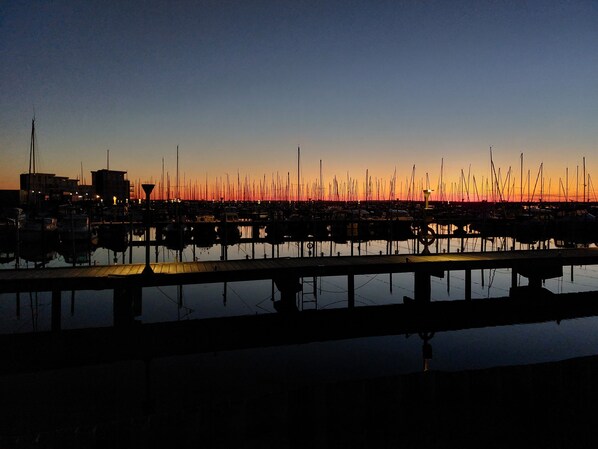 Ein Yachthafen bei Sonnenuntergang mit vertäuten Booten, einer Anlegestelle und Gebäuden im Hintergrund.