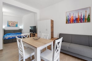 Habitació moderna amb taula de dinar, cadires blanques, un sofà gris, televisió i un armari.