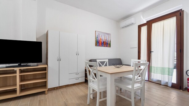A modern room with a TV on a wooden stand, a white dining table with chairs, a white wardrobe, and a colorful painting on the wall. - Apartment 'Casa Leone' with Air Conditioning (Teulada)