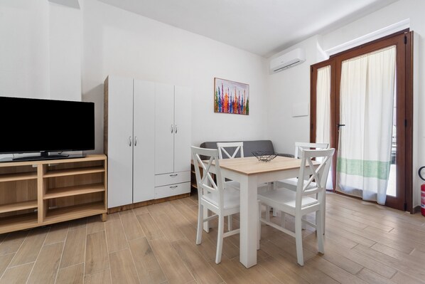 A modern room with a TV on a wooden stand, a white dining table with chairs, a white wardrobe, and a colorful painting on the wall.