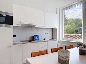 A modern kitchen with white cabinets, a built-in oven, a stainless steel range hood, and a window with a view of greenery. - Stellaross - Koksijde (Koksijde)