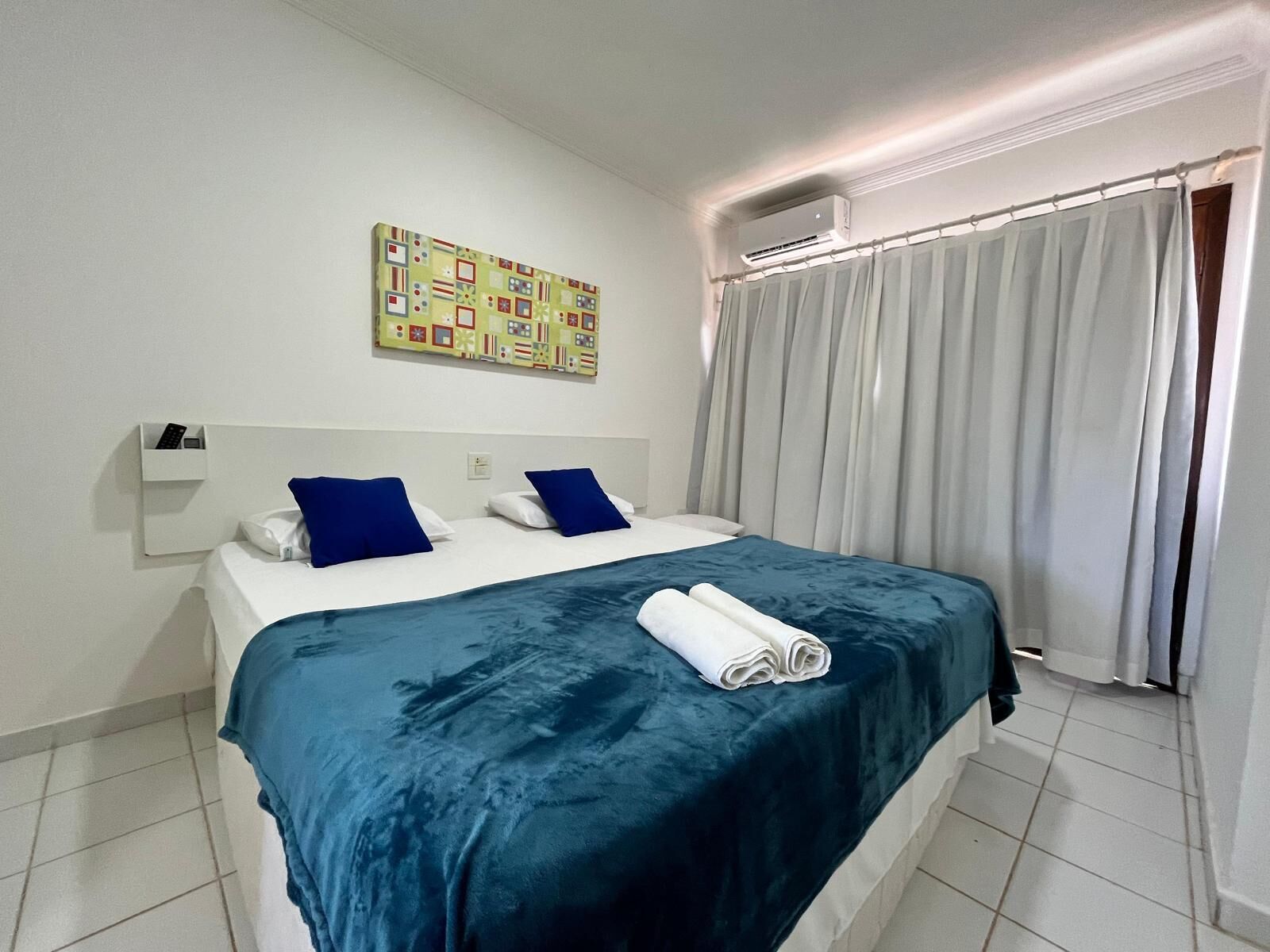 A hotel room with a bed, a wall-mounted air conditioner, a window with curtains, and a colorful artwork on the wall.