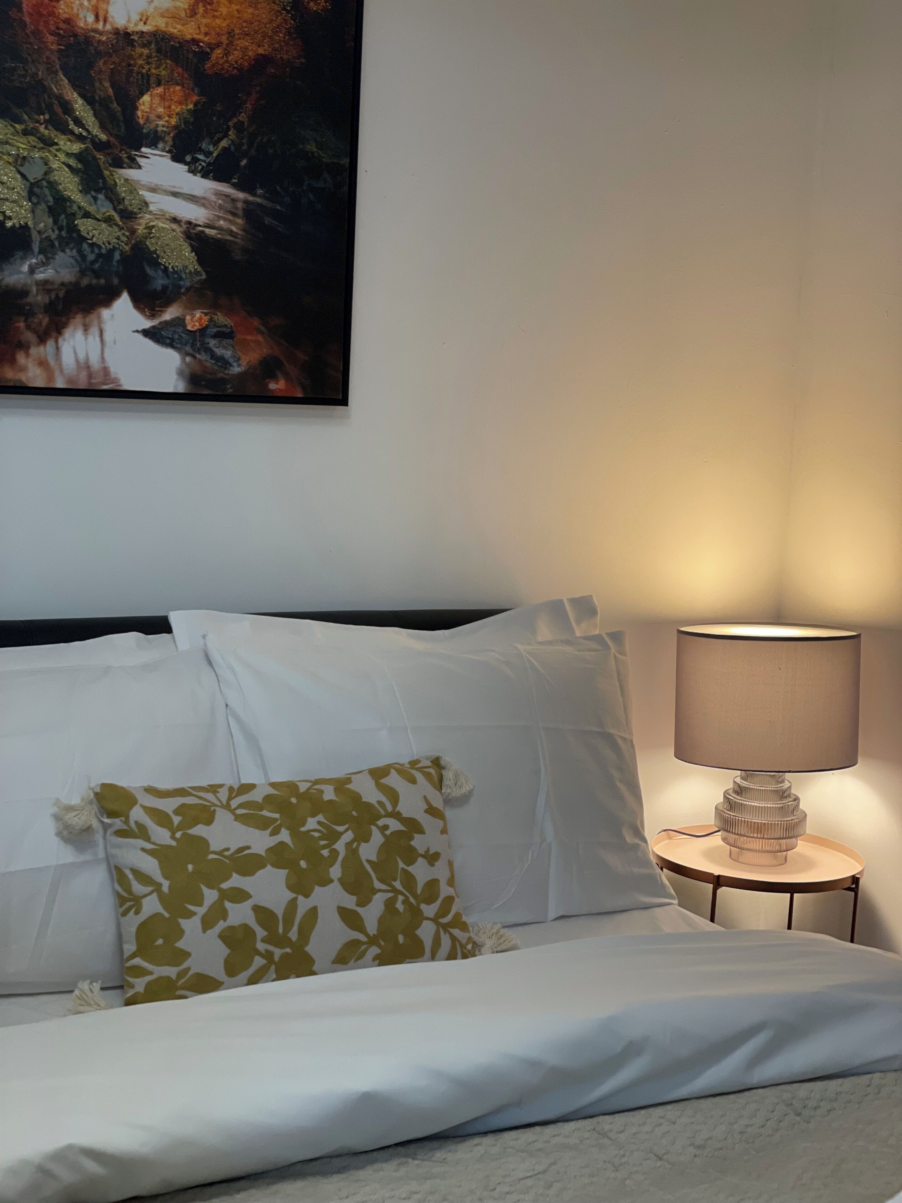 Una cama bien hecha con sábanas blancas, un cojín floral y una lámpara sobre la mesita de noche.