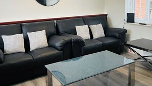 A modern living room with a black leather sofa, a round mirror, a glass coffee table, and a ceiling light fixture.