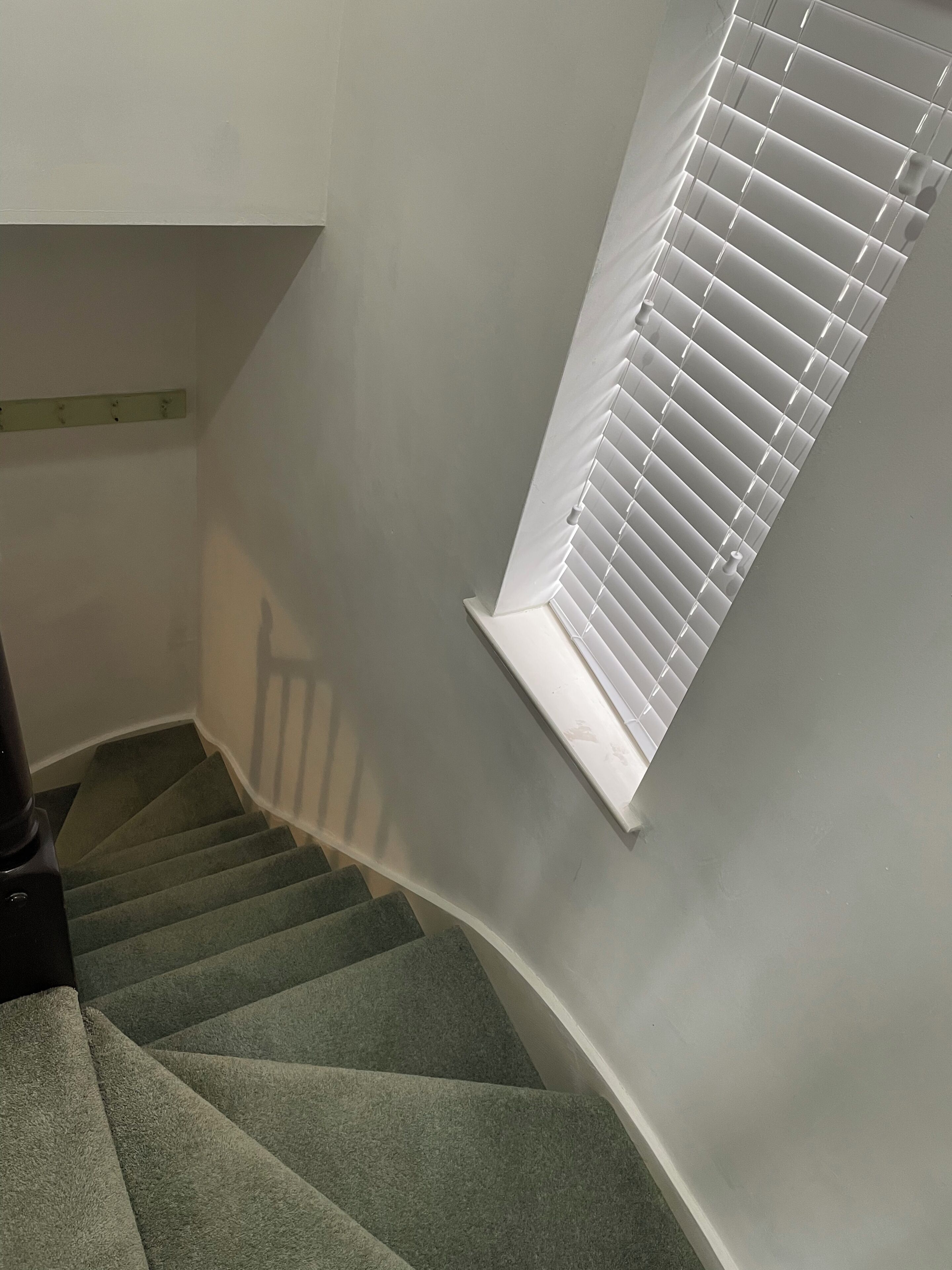 A staircase with carpeted steps leading to a landing, a white wall, and a window with blinds.