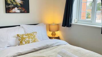 A neatly made bed with white and yellow pillows, a lamp on a bedside table, and a painting of a forest scene on the wall.