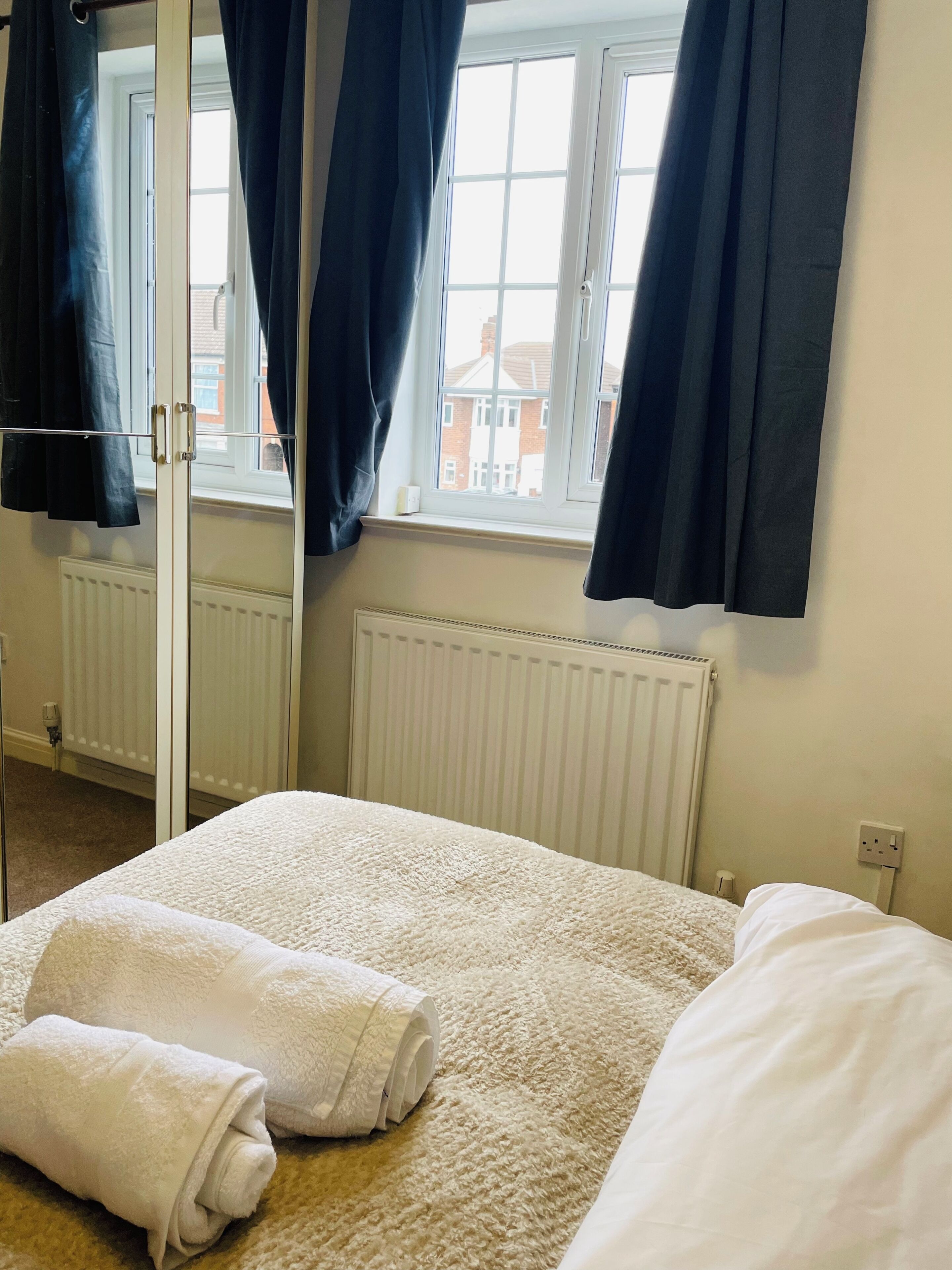 Een slaapkamer met een bed, een radiator, een raam met gordijnen en uitzicht op gebouwen.