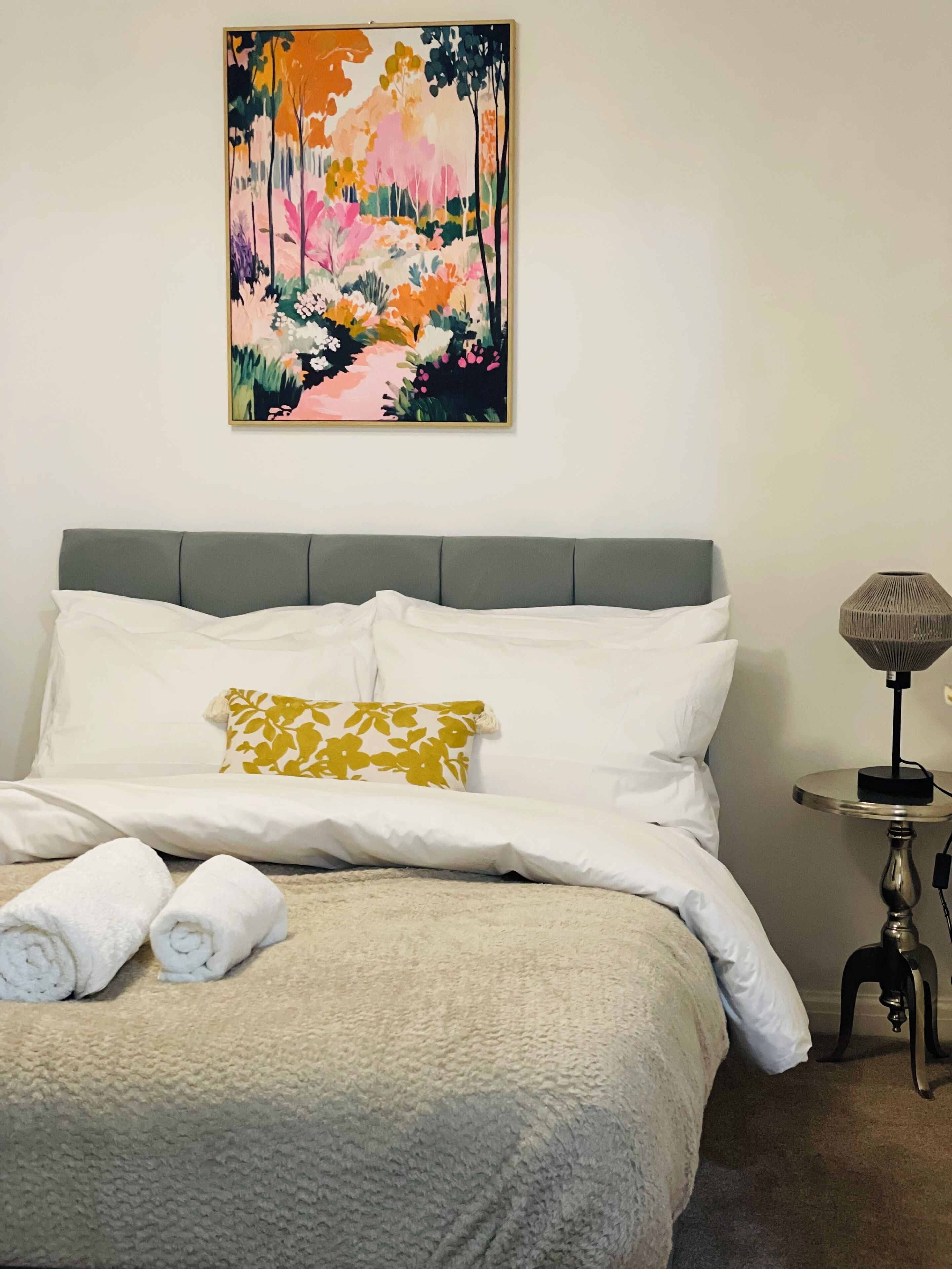 Un letto rifatto con lenzuola bianche e un solo cuscino decorativo, un piccolo tavolino rotondo con una lampada accanto e un quadro astratto dai colori vivaci appeso al muro.