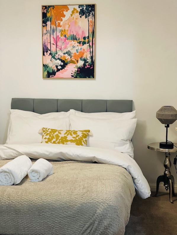 Un lit bien fait, avec du linge de lit blanc et un seul coussin décoratif, une petite table ronde avec une lampe à côté, et un tableau abstrait aux couleurs vives accroché au mur.