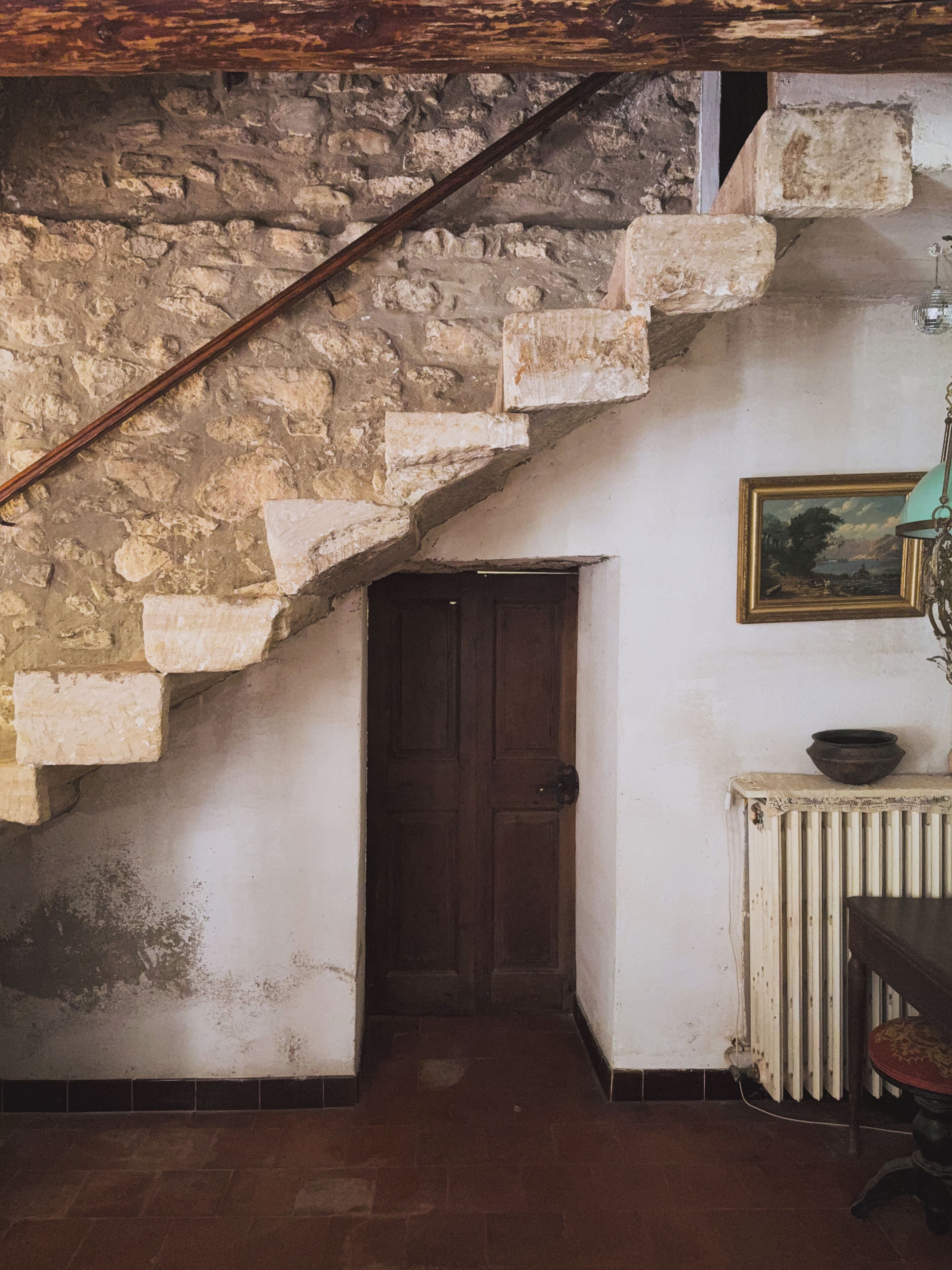 Un escalier en pierre menant à une porte sombre, un radiateur et un tableau encadré accroché au mur.