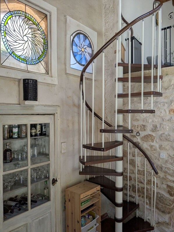 Un escalier en colimaçon avec des marches en bois et des rampes blanches, une vitrine contenant des bouteilles et deux vitraux.