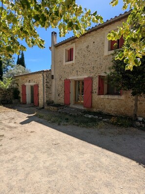 Un bâtiment en pierre avec des volets rouges, un chemin de terre et un ciel bleu azur.
