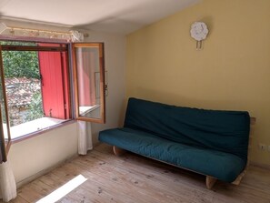 Una habitación con piso de madera, un sofá verde, una ventana con cortinas y un reloj de pared.