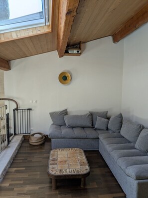 Un salon moderne avec un canapé d’angle gris, une table basse en bois surmontée d’un plateau en mosaïque, une petite table ronde avec un projecteur, et un plafond en bois avec des poutres apparentes.