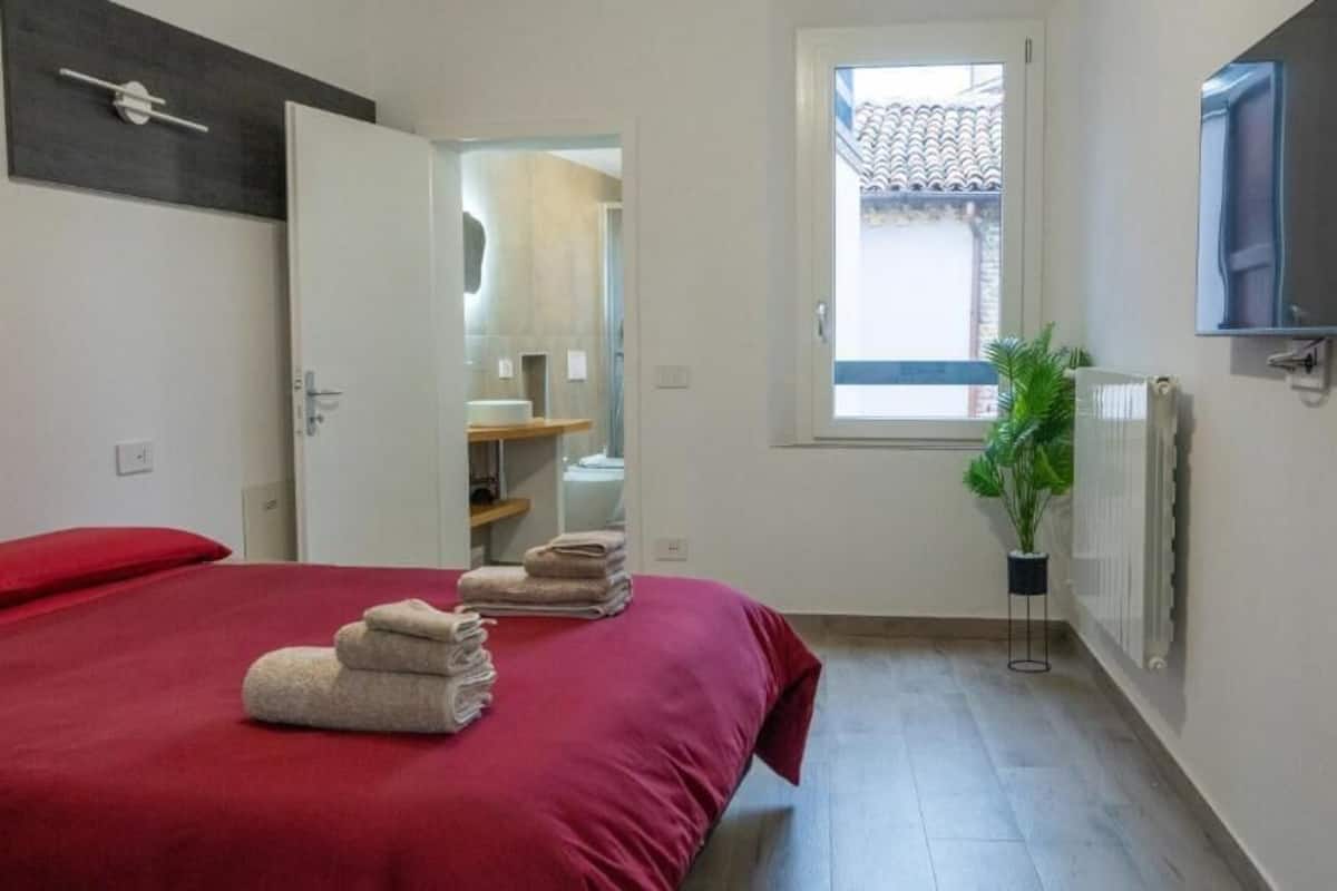 A bedroom with a bed, towels, a window, a radiator, and a TV.