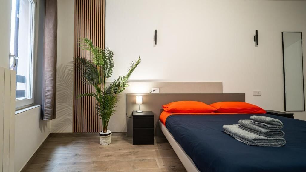 Une chambre à coucher avec un lit, une table de chevet, une lampe, une plante en pot et une fenêtre.