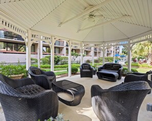 Ein überdachter Außenbereich mit Korbmöbeln, einem Deckenventilator und Blick auf ein Gebäude mit Balkonen.