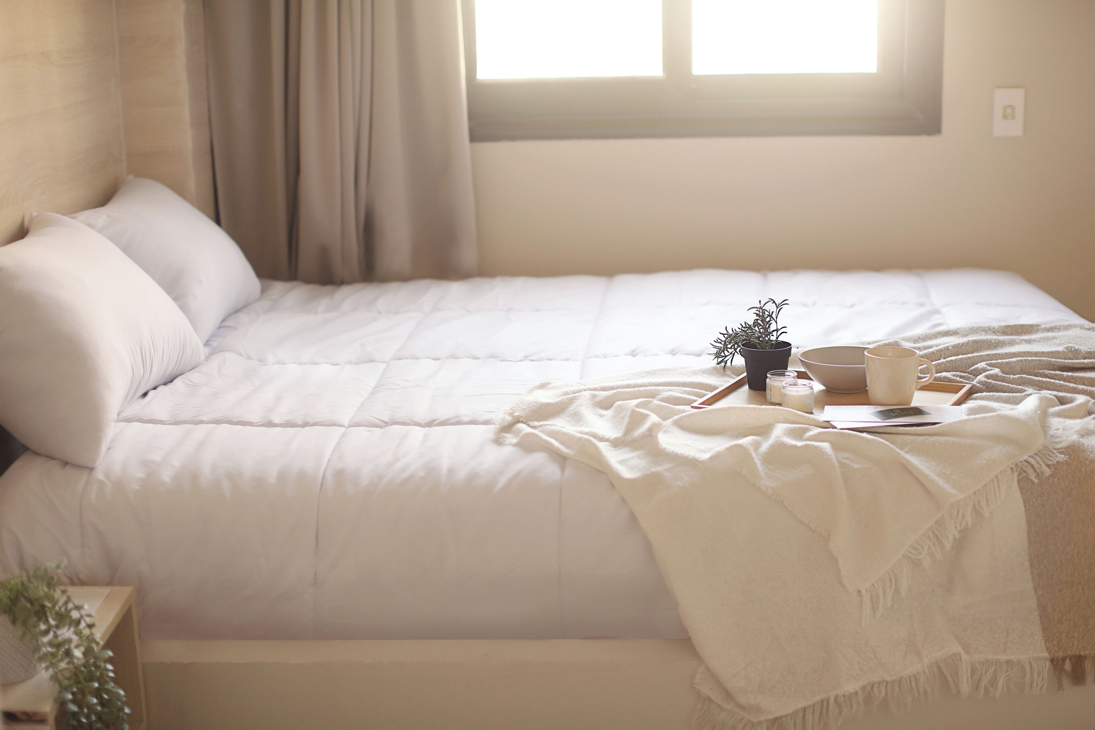 A neatly made bed with a white comforter, a small potted plant, and a tray with candles and a book.