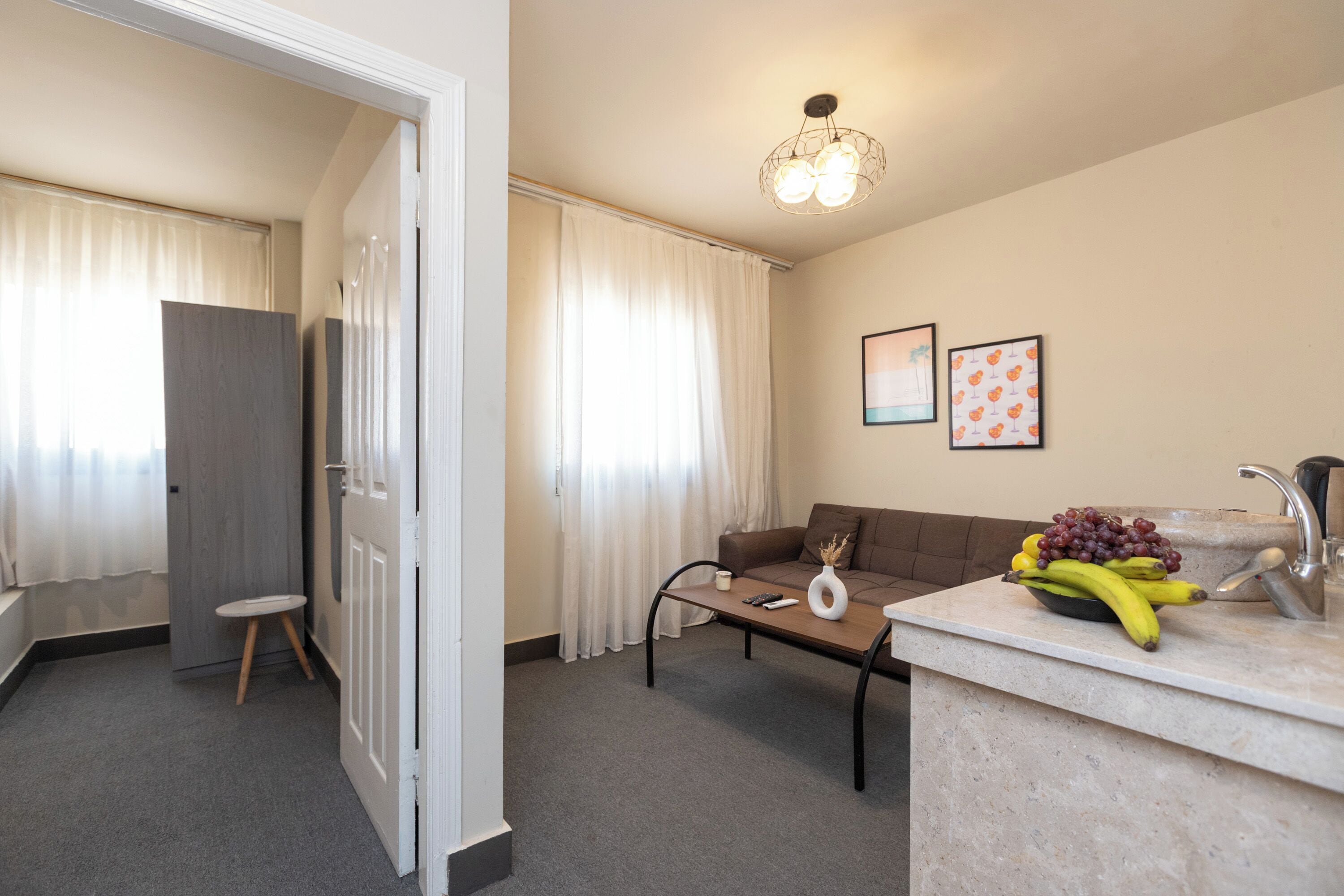 A modern hotel room with a sofa, a coffee table, a small stool, a fruit bowl, and a sink with a faucet.