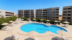 A swimming pool surrounded by lounge chairs and umbrellas, with a backdrop of multi-story buildings. - Seven Stays Sidi Abd El-Rahman (El Alamein)